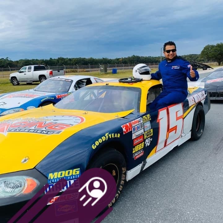 Man in blue racing suit gives thumbs up on a yellow number 15 race car.